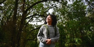 a woman with brown skin and dark curly hair in a gray sweatshirt looks happily at the trees around her while on a walk outdoors