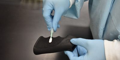 a black gun handle is swabbed by a person wearing light blue gloves and a lab coat