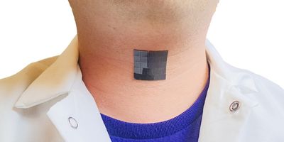 Photo of a man’s neck with a black adhesive square attached outside the throat, the man is wearing a blue t-shirt and a white lab coat, with a white background